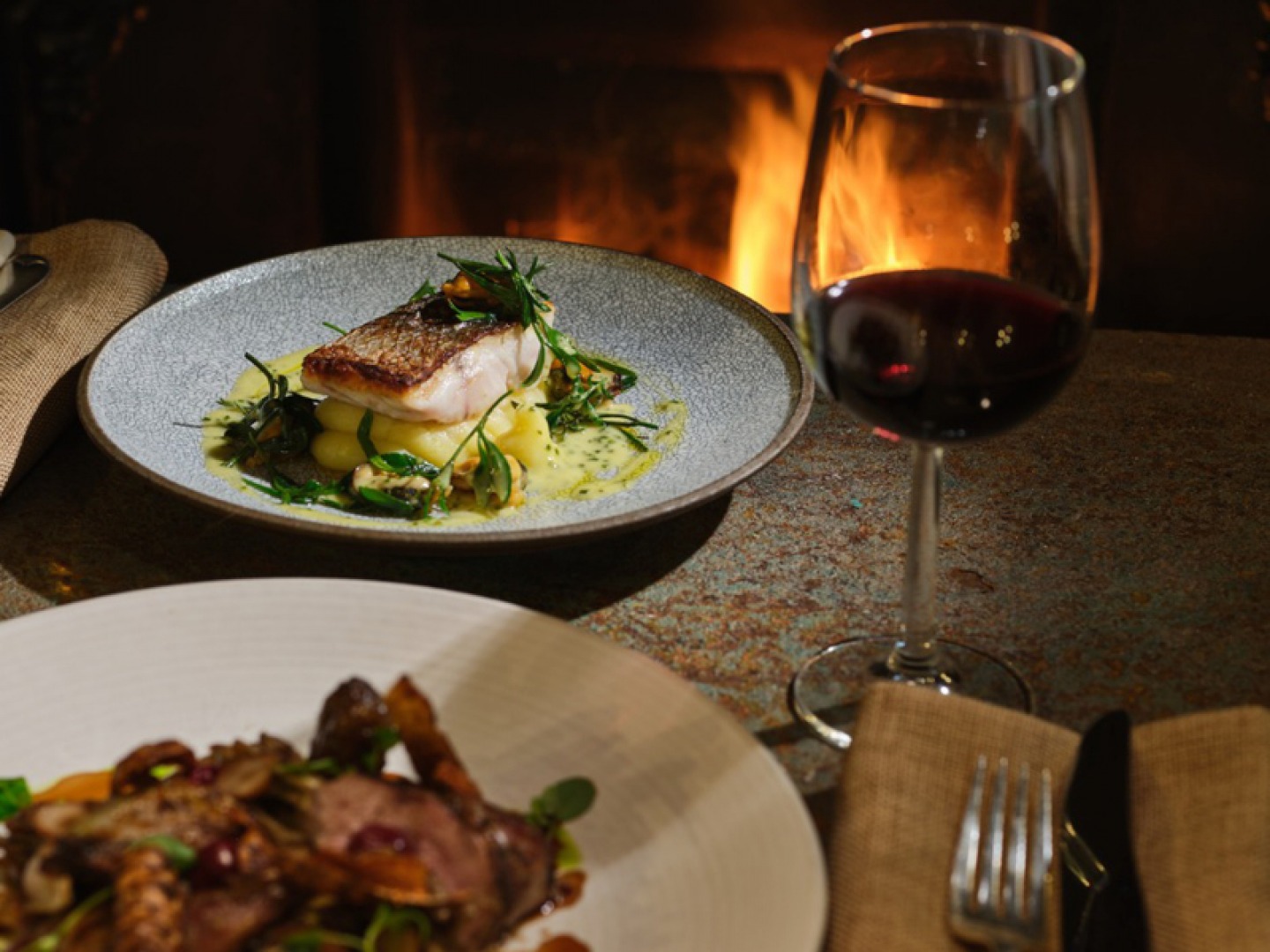 two plates of food in front of a fire with a single glass of red wine