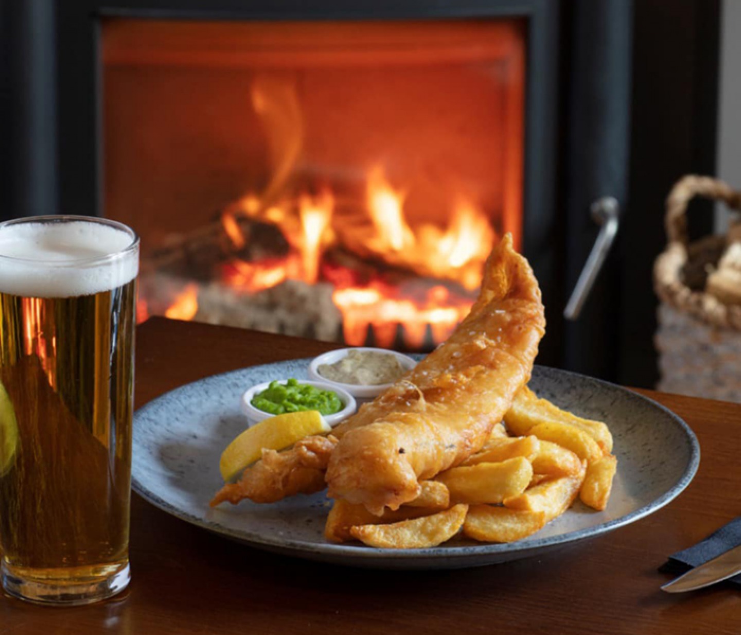 a pint of beer with fish and chips in front of a fire