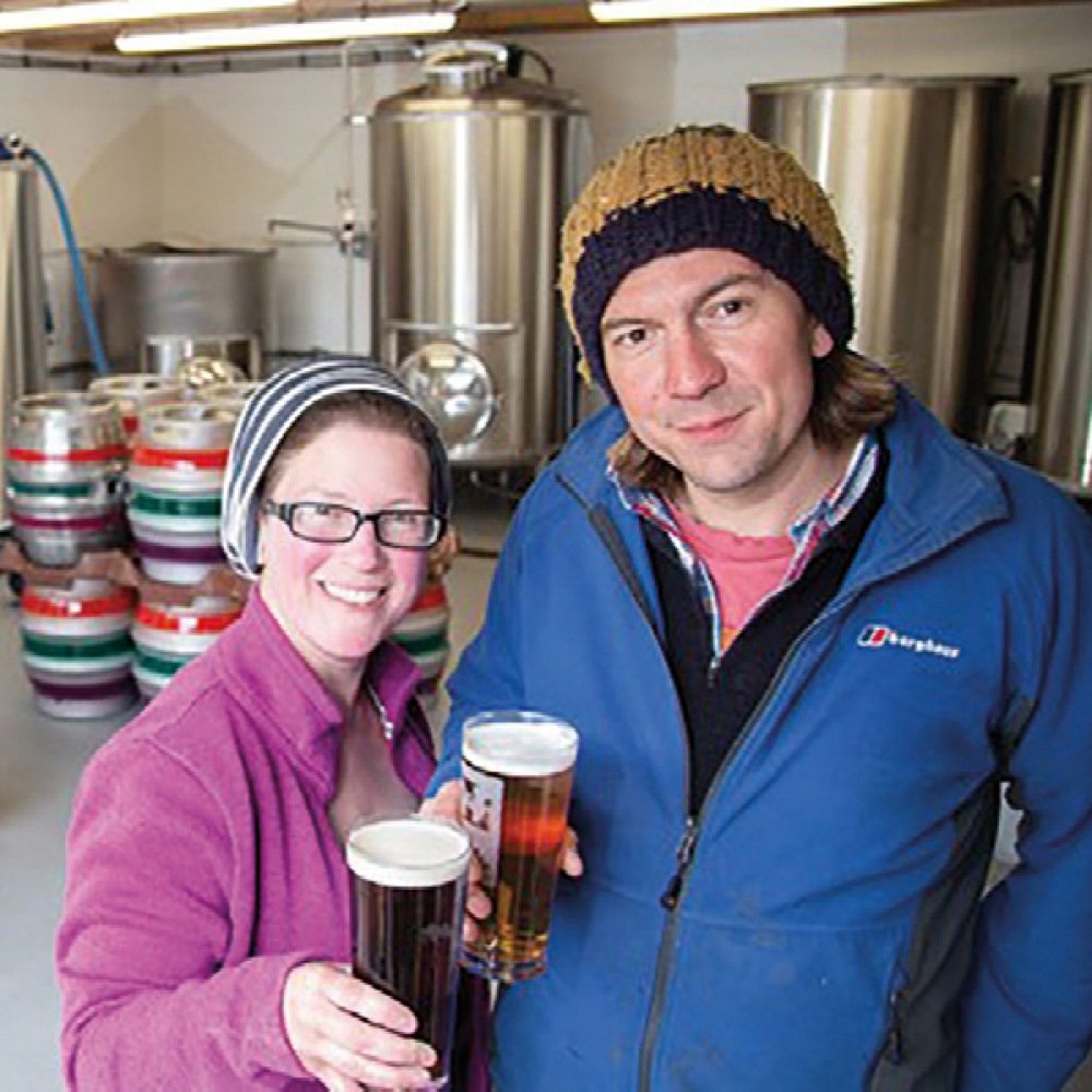 man and woman each holding up a pint inside a brewery