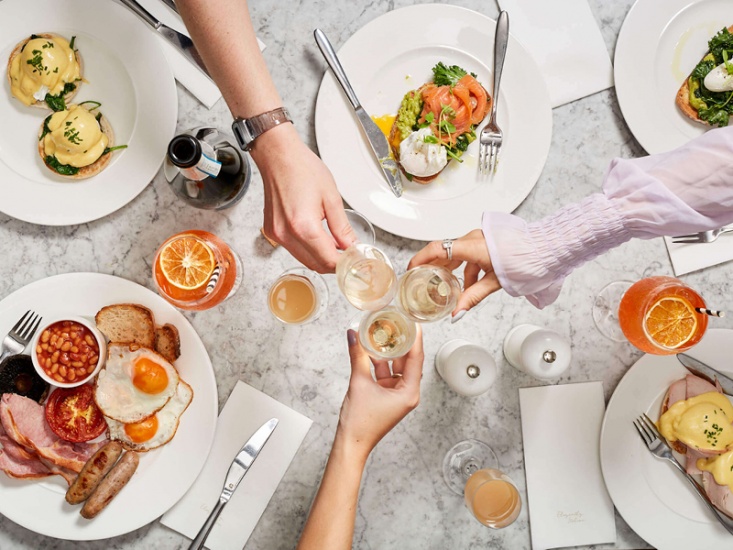 3 three people with glasses of champagne cheering' in the middle of the table with a selection of breakfast on white plated underneath