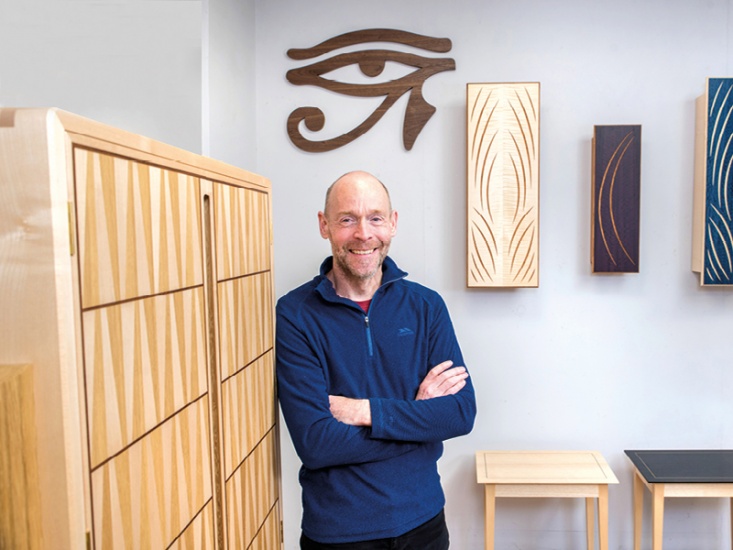 man smiling in a blue jumper leaning against a wooden cabinet