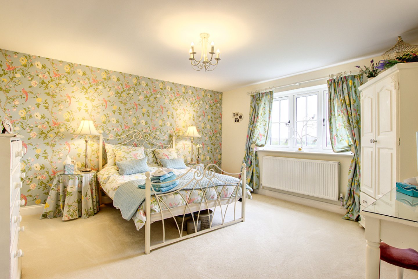 Cottage style bedroom with white metal bed and floral wall paper and soft furnishings