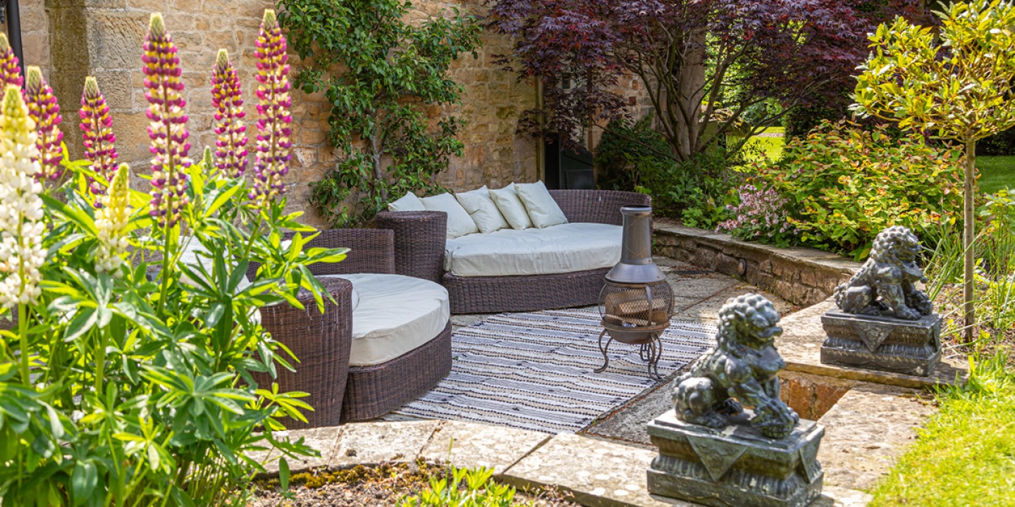 Patio surrounded by plants and foliage with large wicker and cushion seats, steps leading up to 2 lion statues