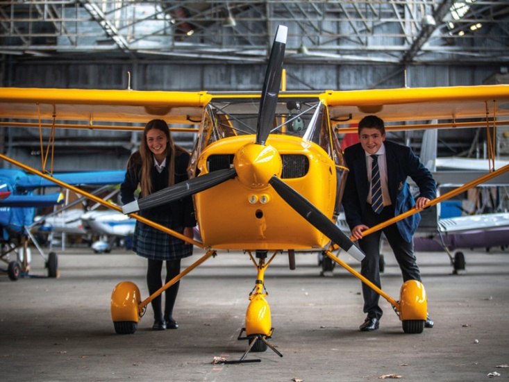 two students crouching under the wings of a yellow propeller plane