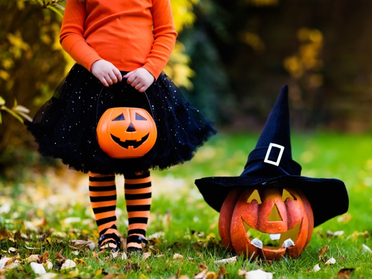 Gril dressed for halloween holding a pumpkin bucket standing next to a pumpkin wearing a witches house