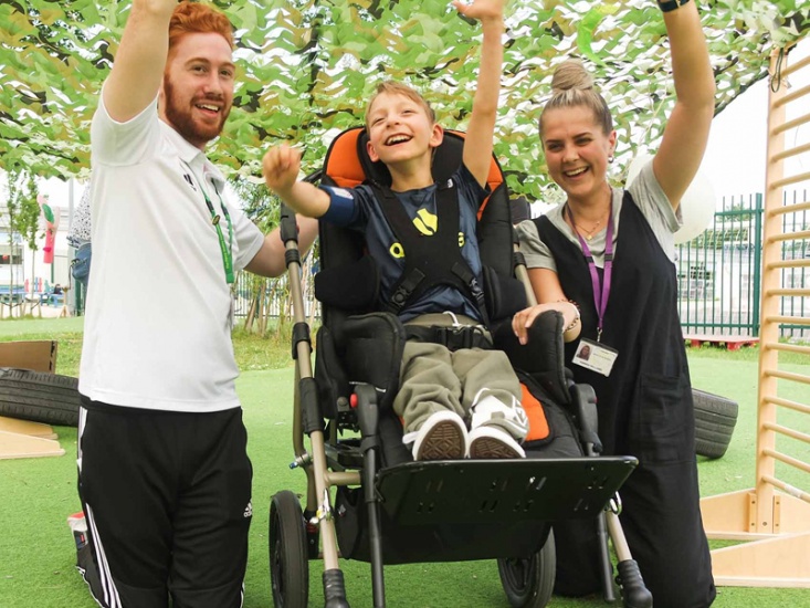 child in a wheel chair in between 2 staff from Percy Headley Foundation all cheering with their arms in the air