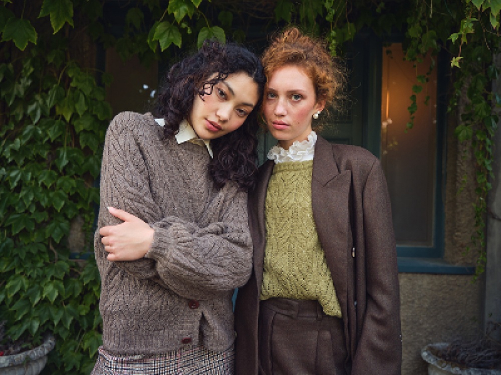 2 models wearing wooly jumpers in front of a window of foliage