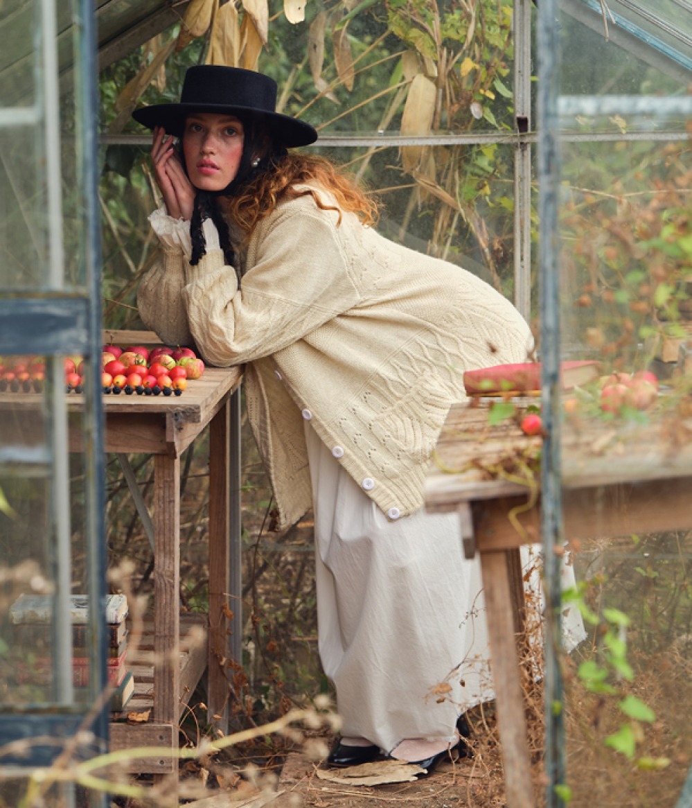 woman in a green hose wearing a cream knitted cardigan and black hat