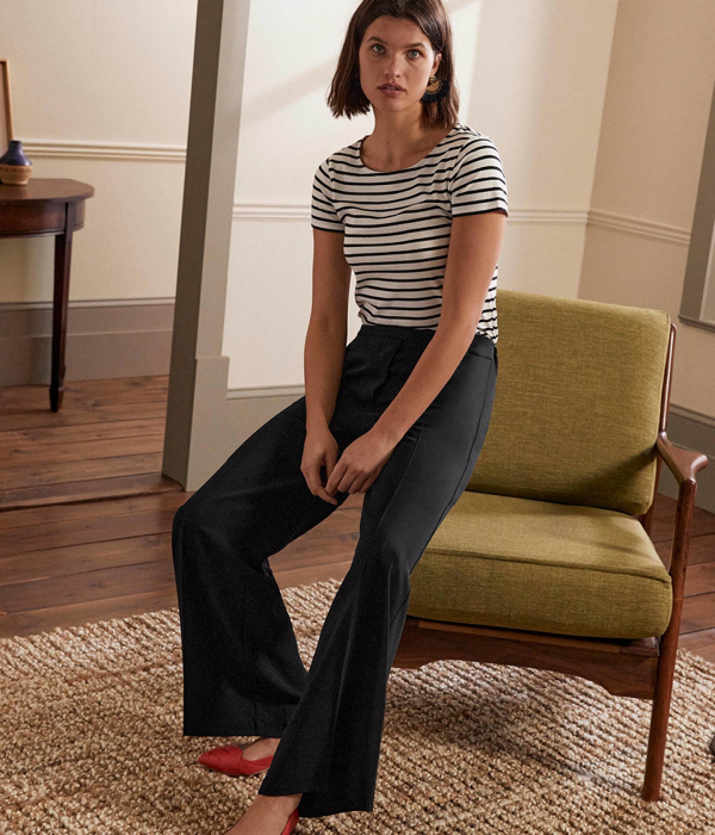 model sitting on the arm of a green chair wearing wide leg trousers a striped T shirt and red pumps