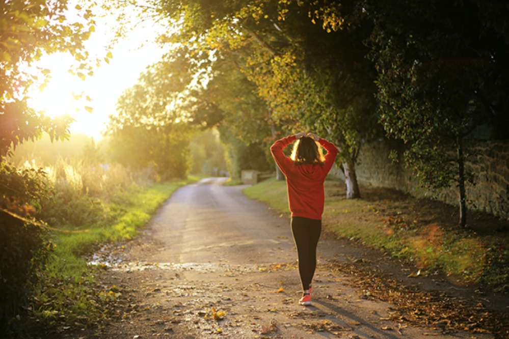 woman walking along a tree lined lane with hands on head in autumn