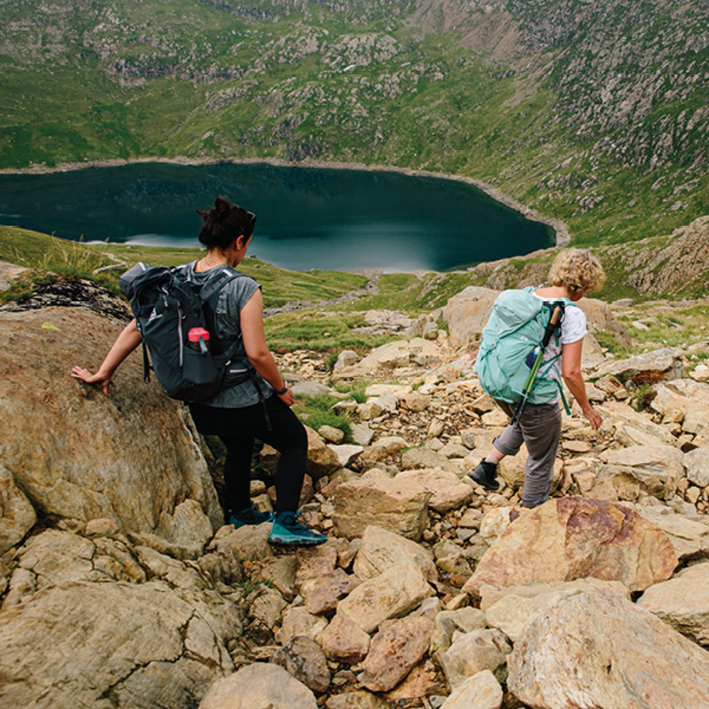 2 women climbing down a mountain side with a tarn in the lower background