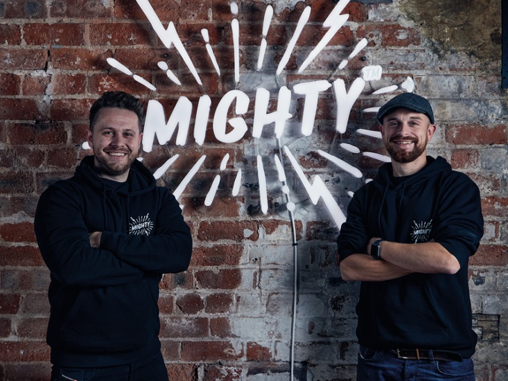 2 men smiling against a brick wall with the word 'Mighty'