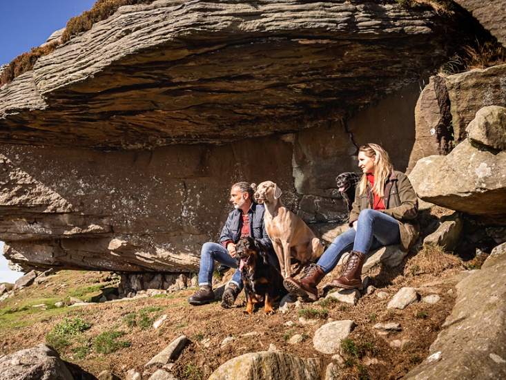 man and woman sat next to 2 dogs under a rock