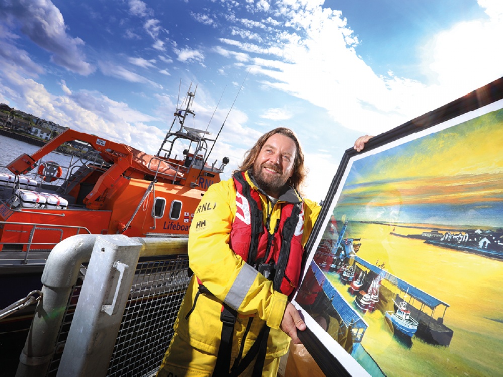 Mark at the harbour dressed in RNLI uniform holding one of his paints of the harbour