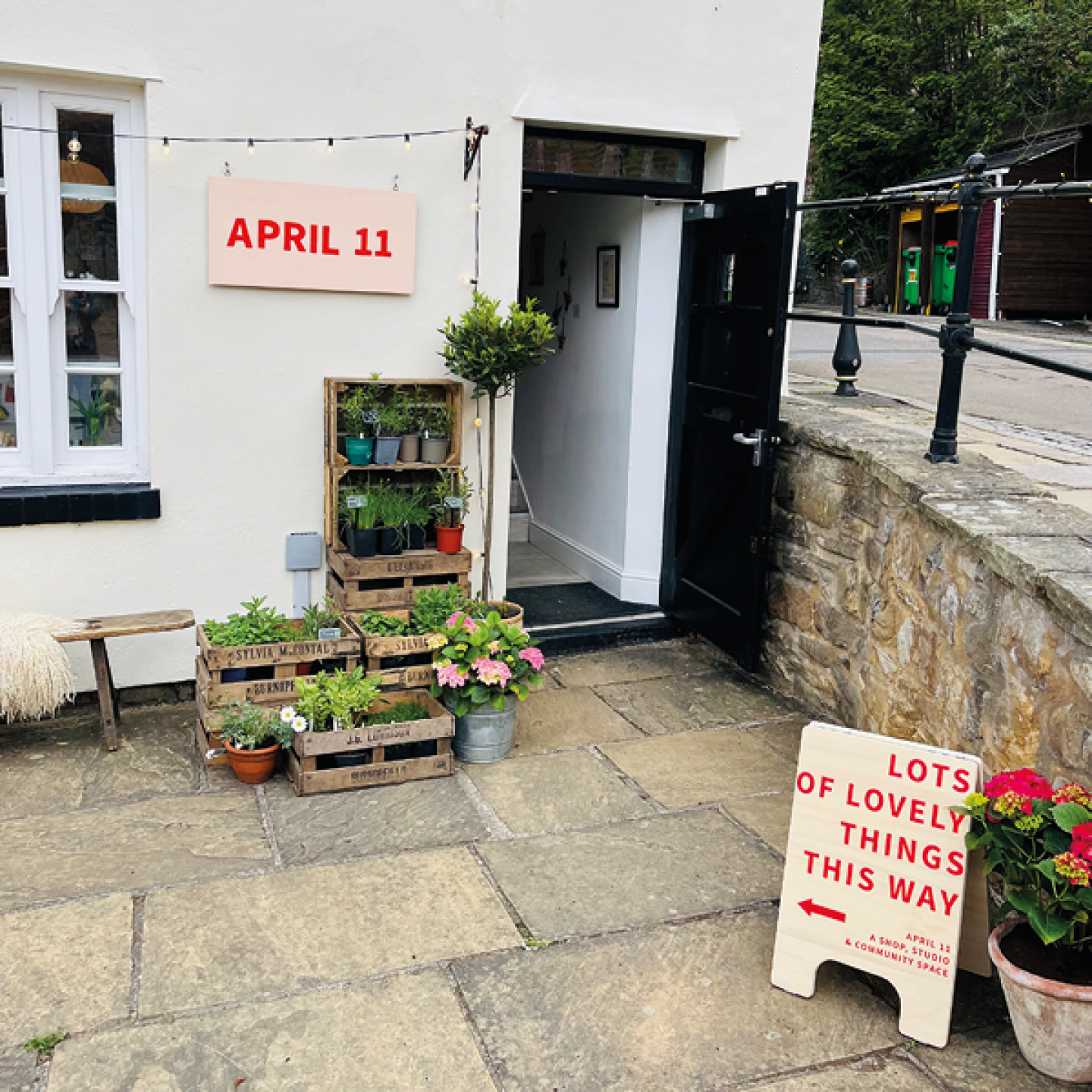Shop front of April 11, with a black door, plants in wooden crates and fairy lights