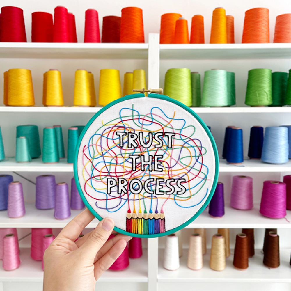 threads on a shelf and a hoop being held up with Trust the Process embroidered into the fabric