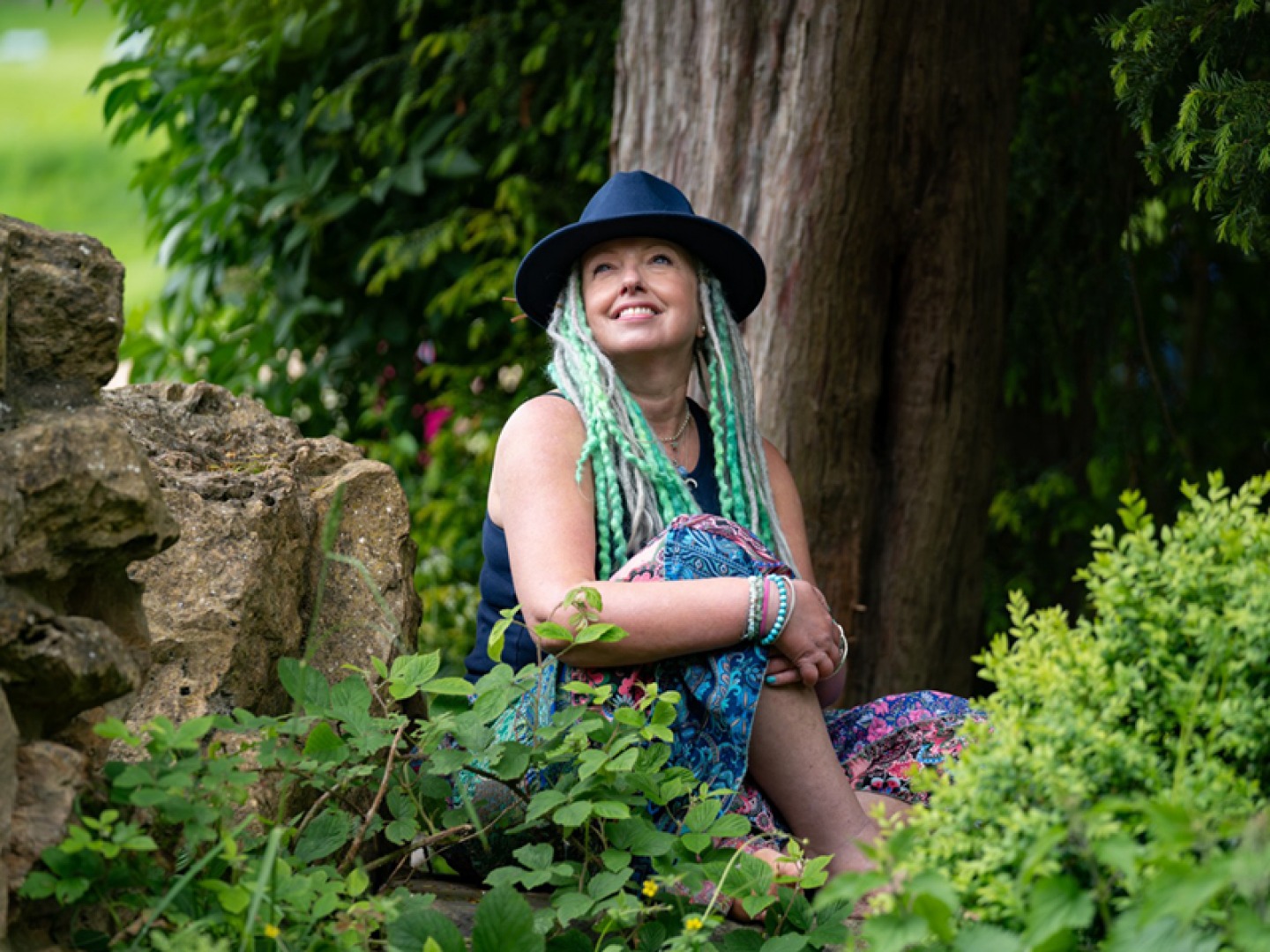 Portrait of a women with green dreadlocks and a black hat sitting on a rock around greenery
