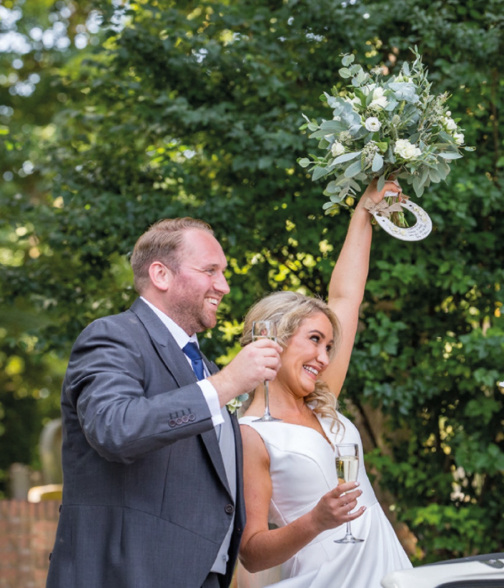 Bride and Groom posing on wedding day with a glass of fizz while bride hold up her bouquet