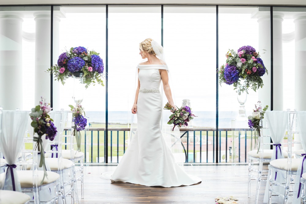 Bride standing in front of picture window looking out to sea with flowers and chairs in the foregorund