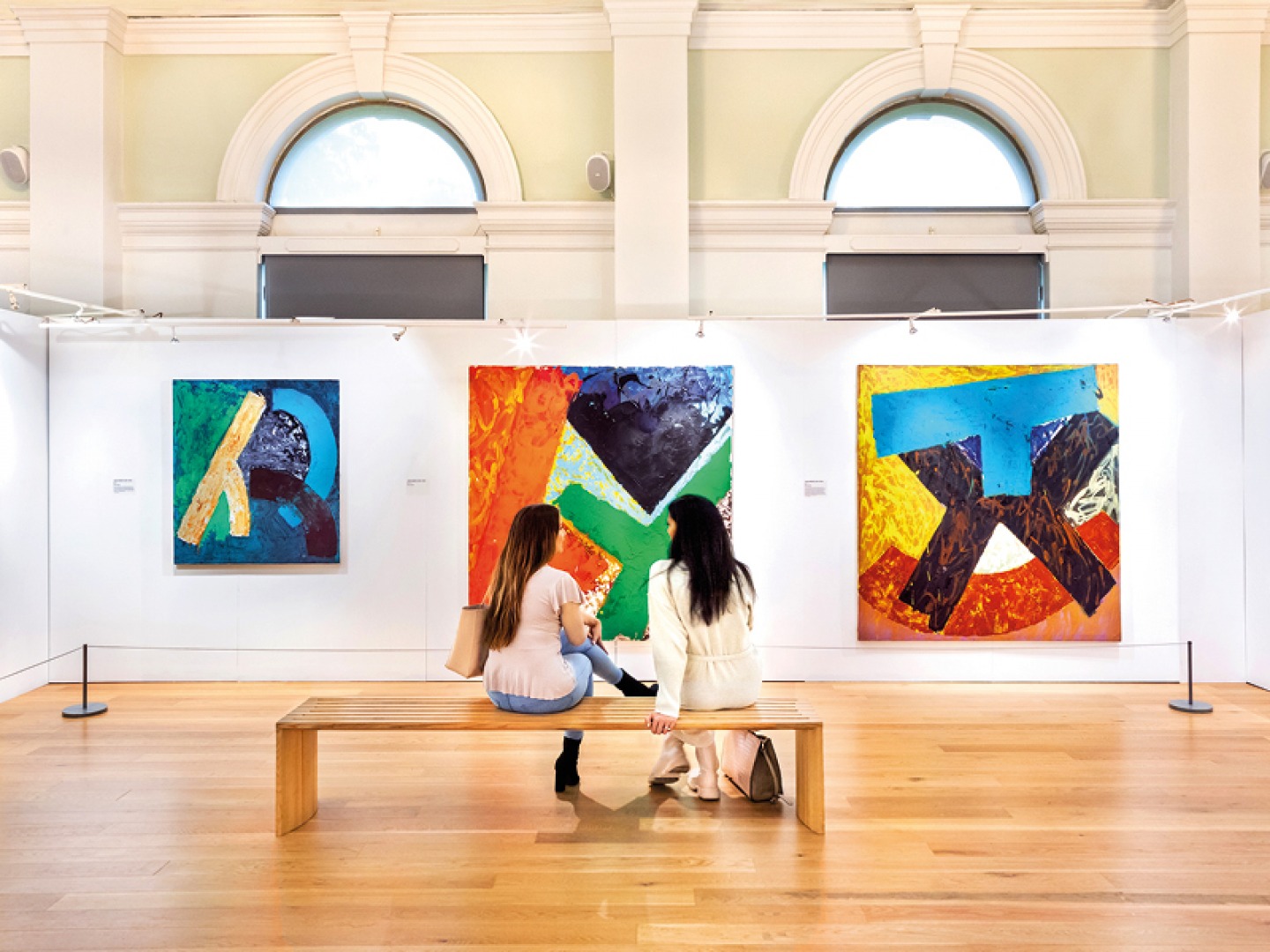 2 people sat on a bench surrounded by paintings in an art gallery