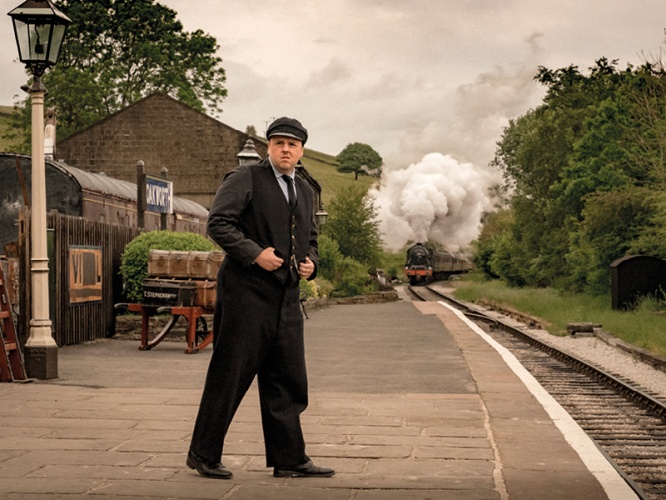 Steam train coming in to a station with guard standing on platform