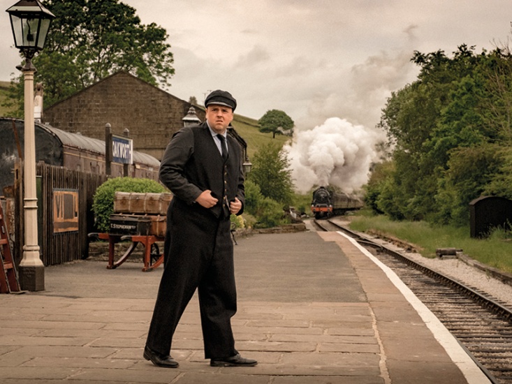 Steam train coming in to a station with guard standing on platform