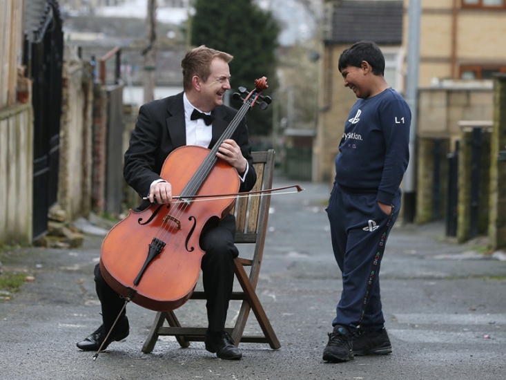 Ben Crick sitting on a wooden chair holding his double base, chatting to a child in a terraced back lane in Bradford