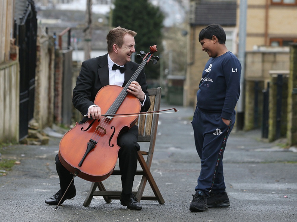 Ben Crick sitting on a wooden chair holding his double base, chatting to a child in a terraced back lane in Bradford