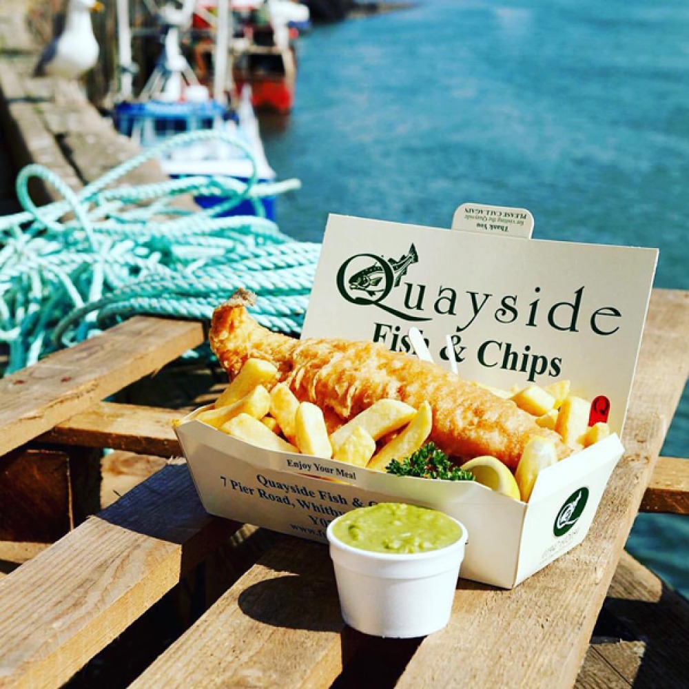 fish and chips in a takeaway box on a wooden table in front of the harbour