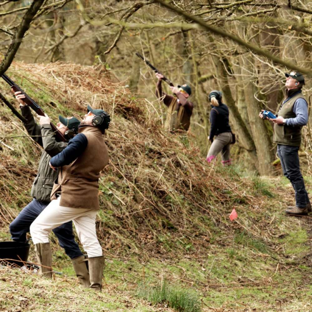 Group of people outdoors in the countryside aiming and shooting shotguns, with grassy fields and trees in the background.