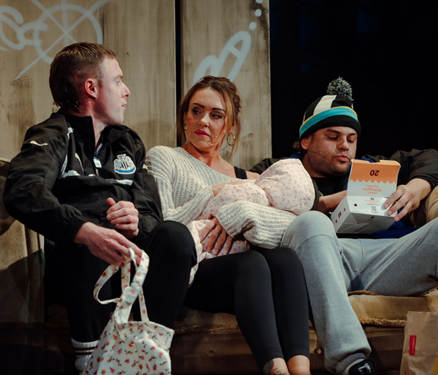 3 cast members sitting on a sofa the woman holding a baby and one male eating chips