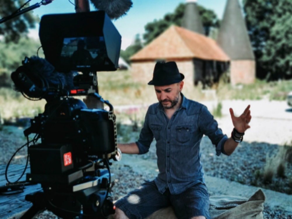 Mark A. C. Brown in front of a big black film camera with a black trilby hat on his head