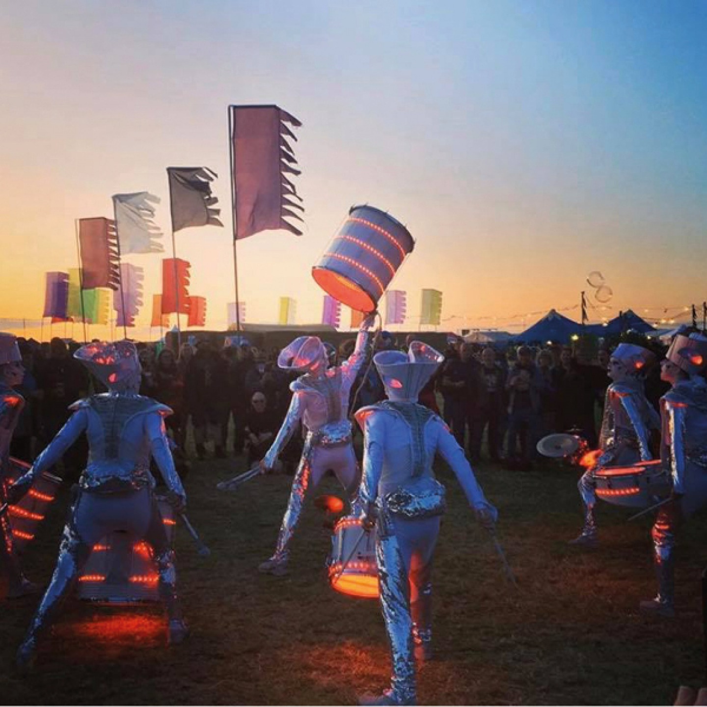 dancers in metallic space suits playing lit up drums at sunset
