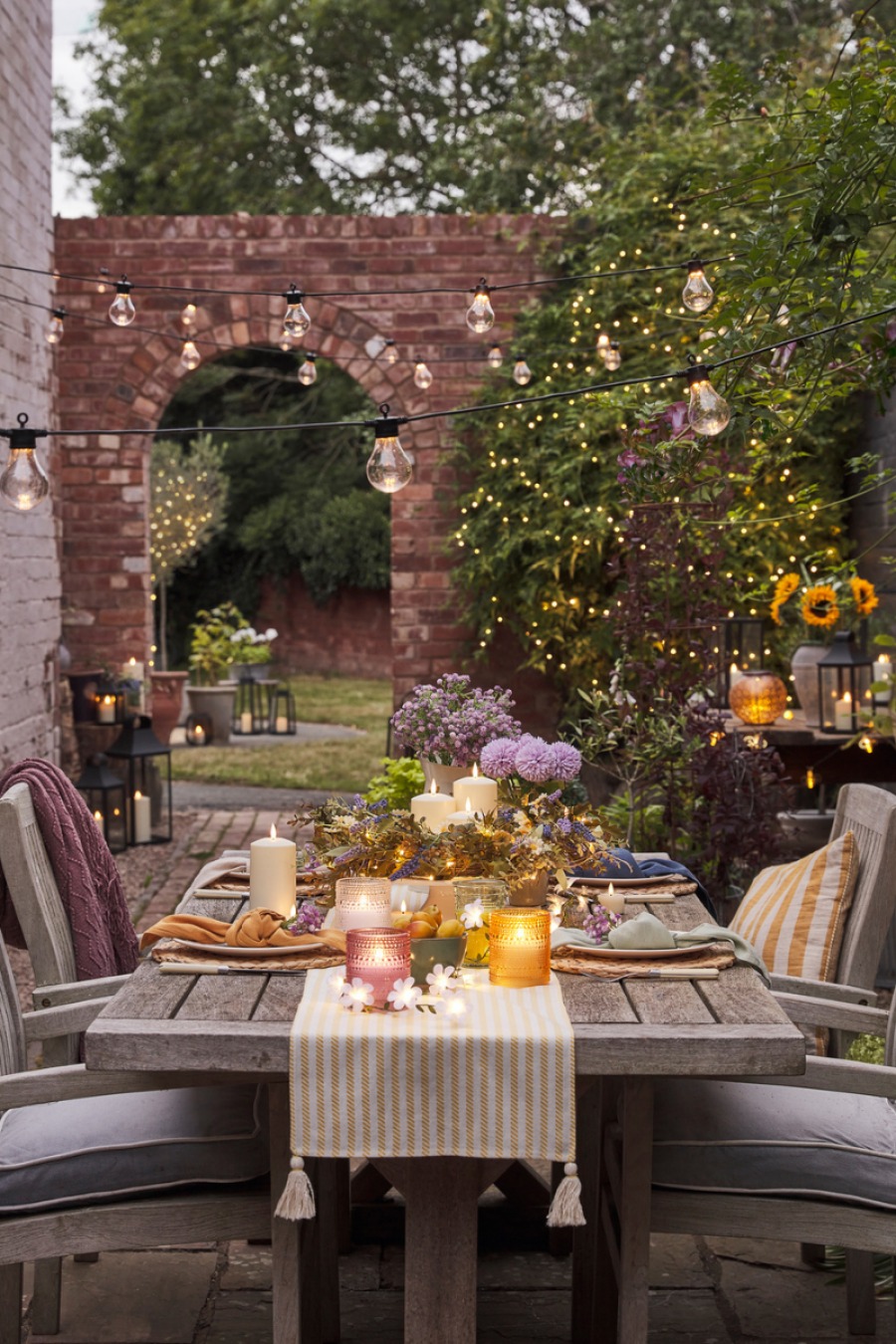 A four person wooden table outdoors with candles a white and yellow runner and flowers on top